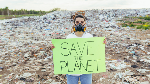 Person in a gas mask in front of a vast landfill, holding a green poster that says "save the planet"
