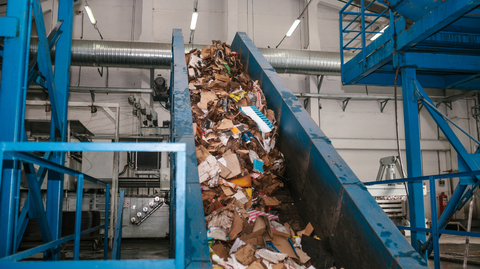 Inside of a recycling facility, large blue metal structures and a conveyor belt with paper and cardboard.