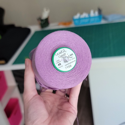 A large cone of a light lilac purple thread, being held in my hand in front of a workstation.