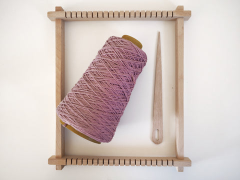 a wooden lap loom with a cone of purple cotton string and a wooden weaving needle