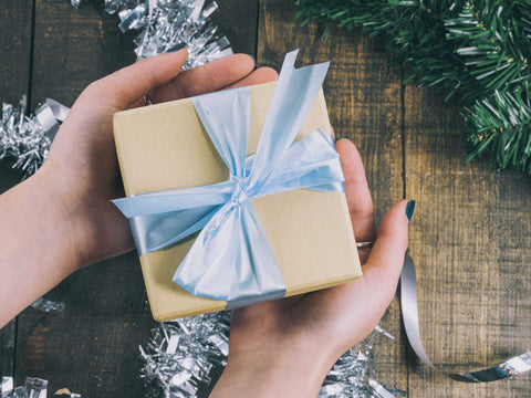 hands holding gift box tied with a blue ribbon with Christmas decorations in background