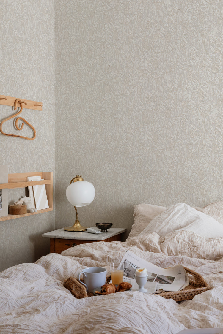 Olof, Floral Pattern Wallpaper in Sand featured on the wall of a bed room, with a bed with soft fabrics and pillows with a side table and an small lamp on it.