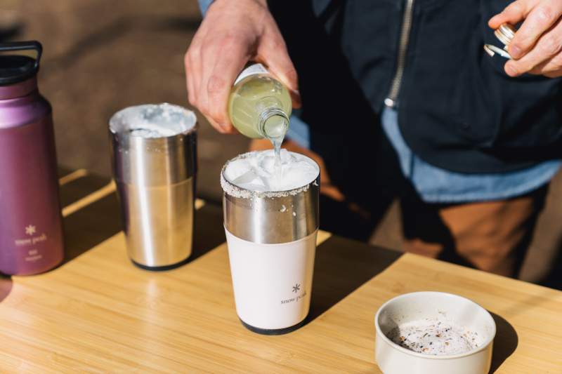 A camper pours sparkling yuzu into a Shimo Tumbler sitting on a bamboo table as they prepare a Yuzu Shandy.