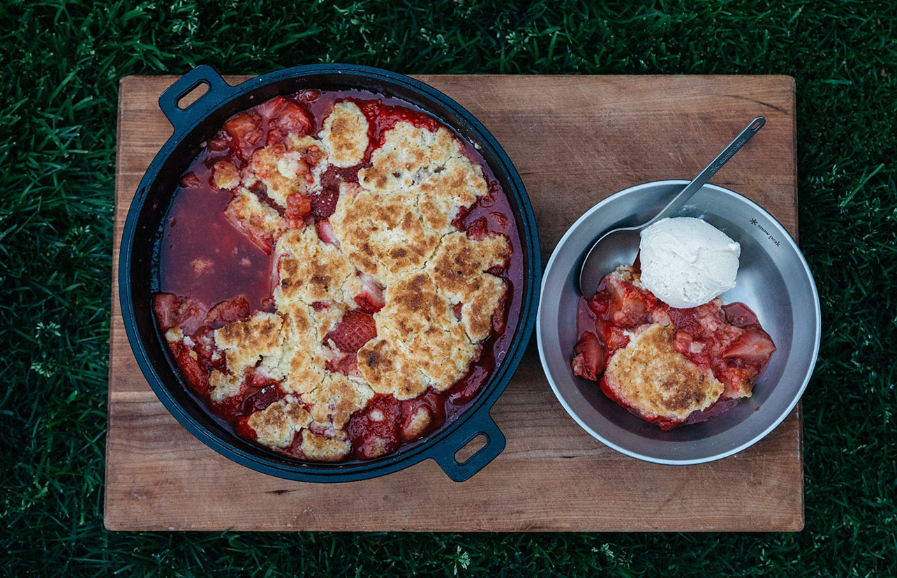 Strawberry Buttermilk Biscuit Cobbler in a cast iron pan with a serving plated into a bowl sitting next to it.