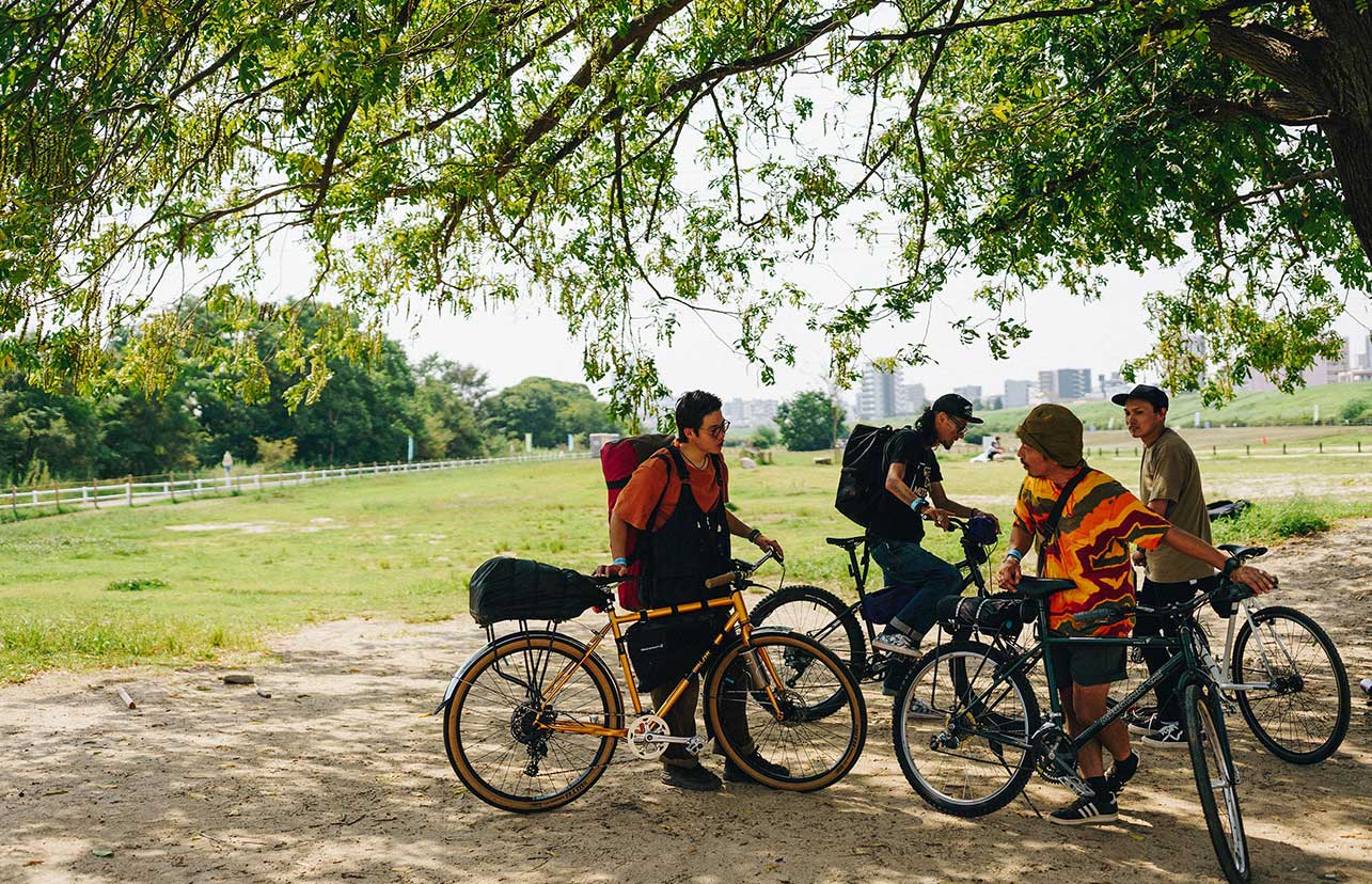 A group of four bike riders prepare to go on a ride in a park. Gear is stored on each of their bikes.