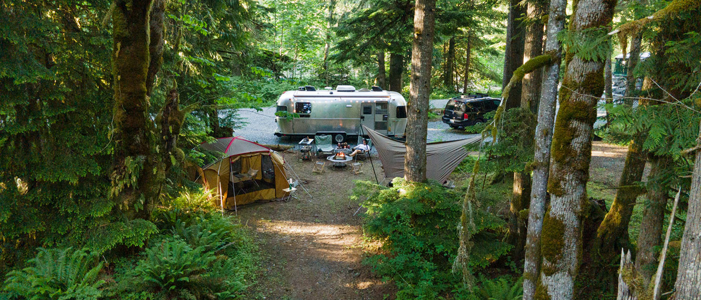 Image of a camp setup with a vintage Airstream, Snow Peak shelter and tarp, and a campfire nestled into the woods of the Pacific Northwest.
