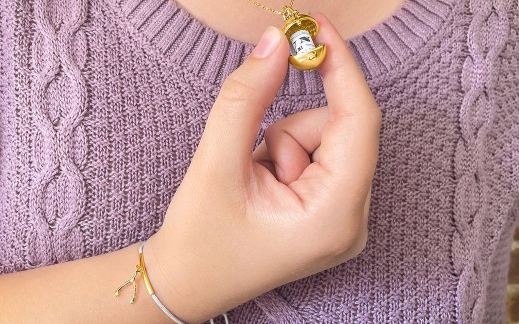 Woman in purple sweater holds Sphere + Wand Locket in between her fingers while wearing Wishbone String Bracelet on her wrist.