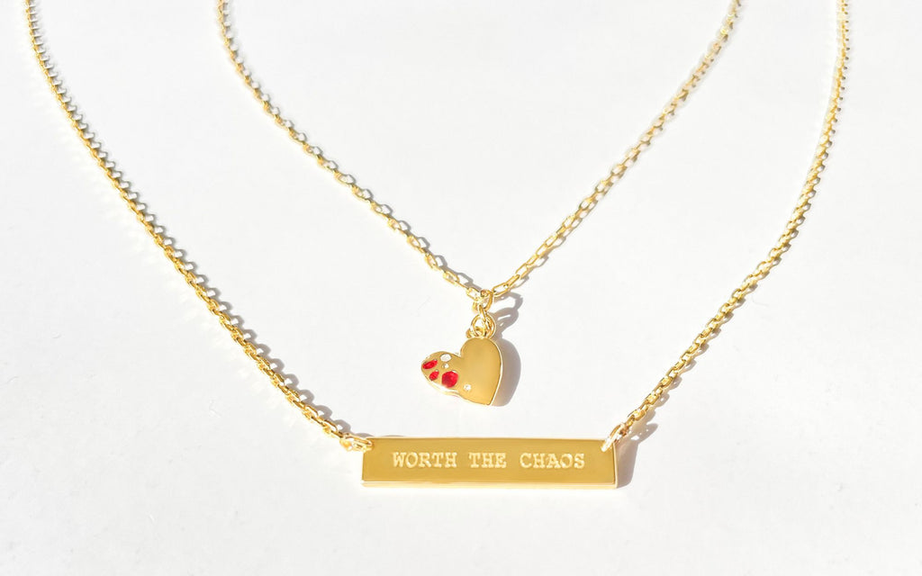 Gold Jeweled Heart Pendant and  “Worth the Chaos” Engraved Bar Pendant lay on white surface.