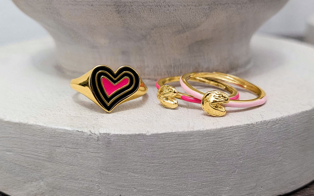 A heart ring beside two fortune cookie rings sitting on top of an off white candle base.