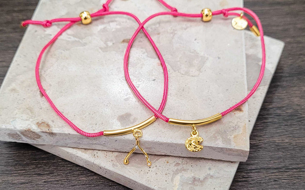 A pink wishbone string bracelet, and a pink fortune cookie bracelet on top of square marble coasters.