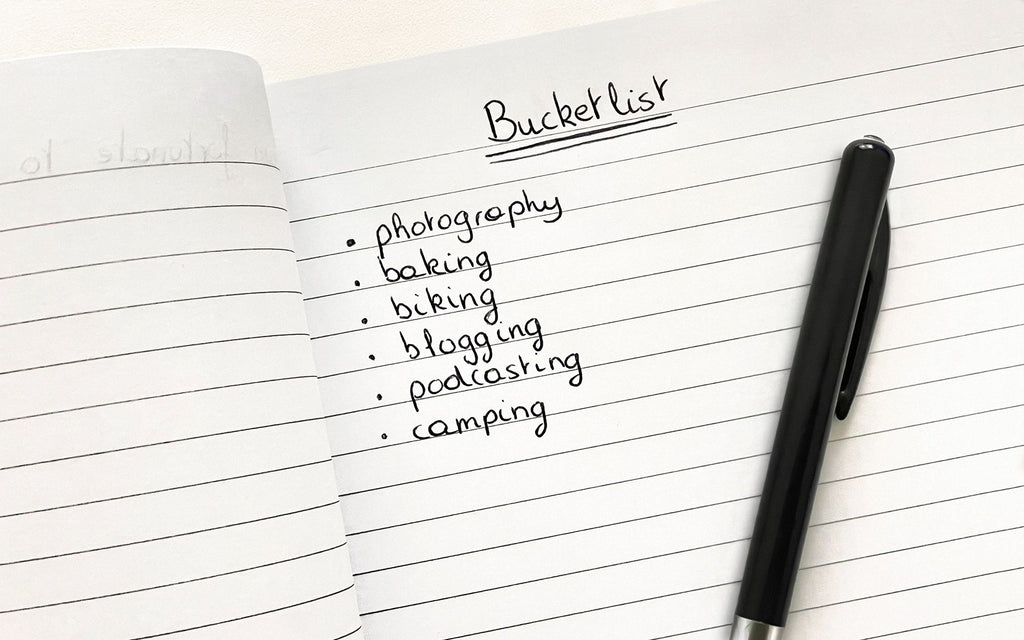 A notebook with the words "Bucket List" written and then bullet points of photography, baking, biking, blogging, podcasting, and camping. A black pen is laying on the notebook paper.