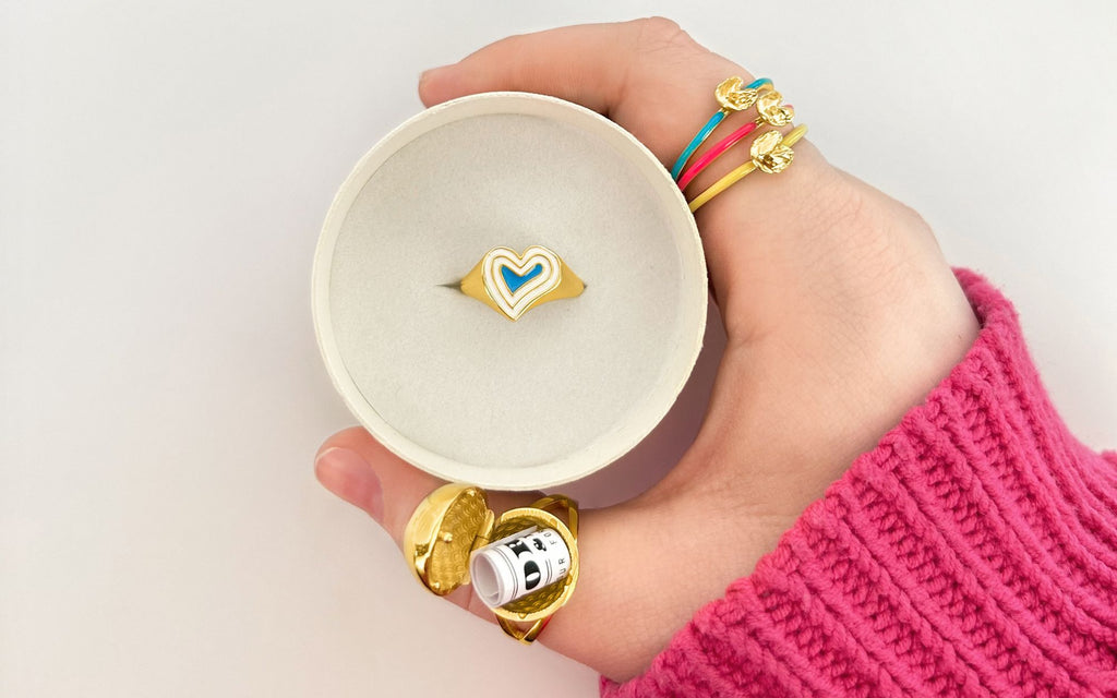 Hand holding a Deco Heart Ring in a ring box while wearing a Gold Sphere Secret RIng and three Fortune Cookie Rings.