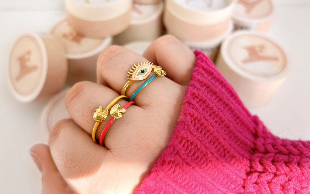 Hand wearing Fortune Cookie RIngs and a Vibrance Evil Eye Ring with ring boxes in the background.