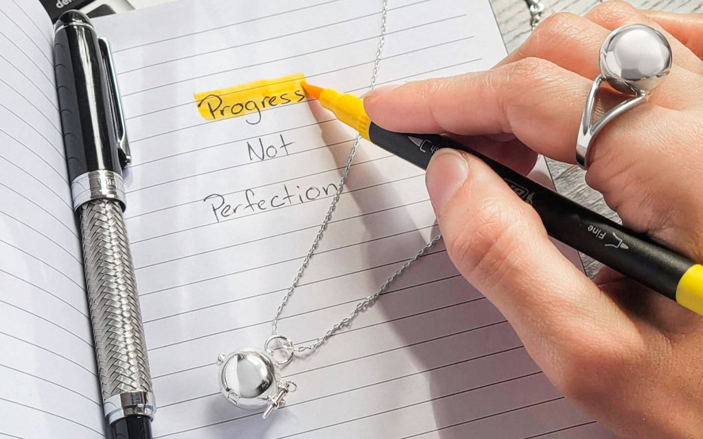 Someone highlighting "Progress NOT Perfection" with a silver Sphere + Wand Locket laying on the notebook and wearing a Silver Sphere Secret Ring 
