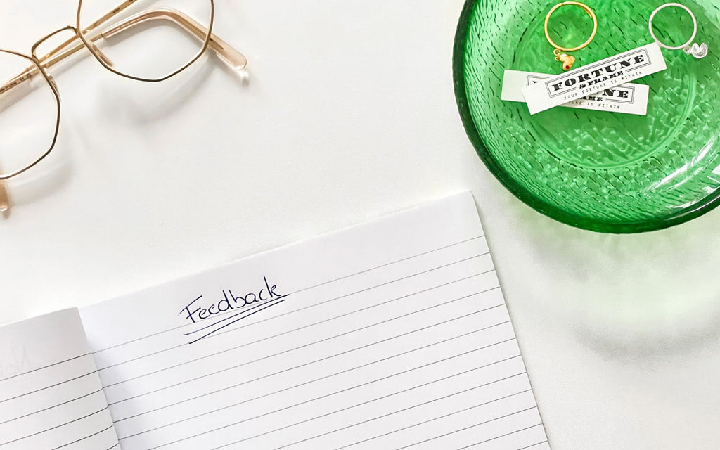 A notebook that reads "Feedback" with glasses and a green bowl above it. In the green bowl are two fortune & frame jewelry pieces along with a gold jeweled heart charm ring and silver fortune cookie charm ring 