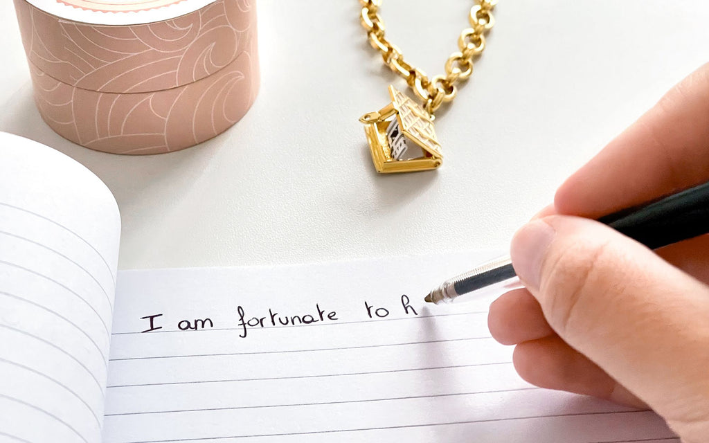 A Fortune & Frame ring box next to a Pearl Secret Diary Book Bracelet that is opened and holding a fortune. In the foreground there is a notebook being written in with the start of an note that says "I am fortunate to h..."