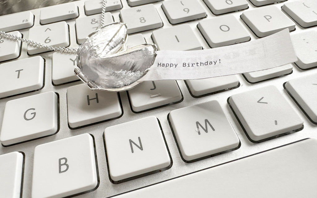 A Silver Fortune Cookie Signature Locket that has a fortune coming out of it that reads Happy Birthday! It is on top of a keyboard. 