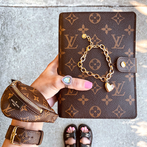 Image of a hand holding a Louis Vuitton planner with the F&F Heart Locket on top. 