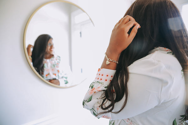 Influencer Natasha Brown looking into a gold circle mirror, fixing her hair with her left hand while wearing the Fortune & Frame Jeweled Heart Bracelet.