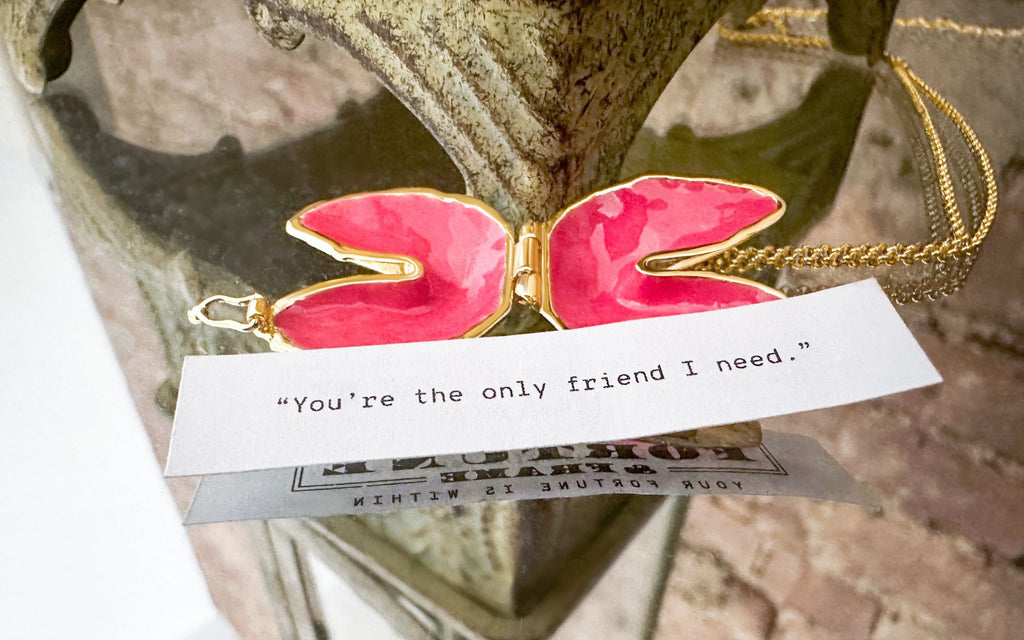 Signature Fortune Cookie Locket (Pink) laying opened on a mirrored table with a fortune in front of it that reads "You're the only friend I need.”