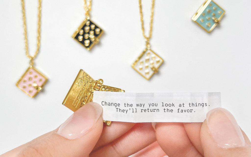 Blush, Black, Ivory, and Turquoise Stardust Locket all lined up in the background with an open locket and a hand holding a fortune. the fortune reads "Change the way you look at things. They'll return the favor." 