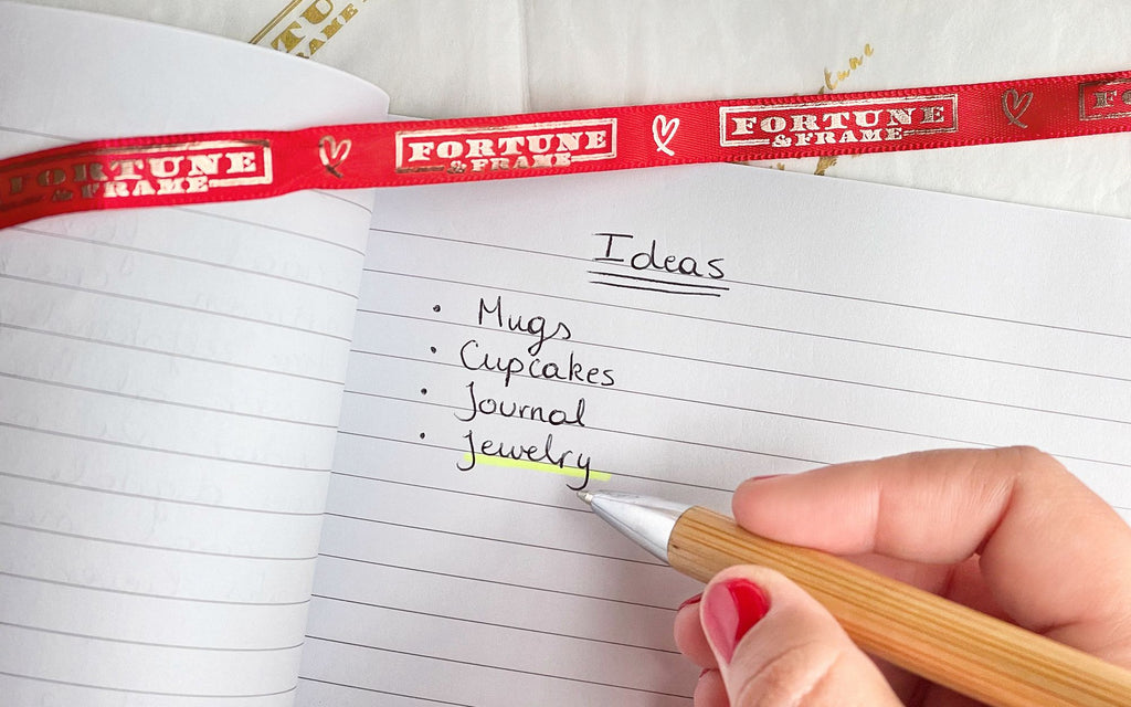 A notebook opened on top of Fortune & Frame tissue paper and someone writing ideas for gifts. The list says Mugs, Cupcakes, Journal, Jewelry. Jewelry is highlighted in yellow. Across the top of the notebook is a red Fortune & Frame ribbon.