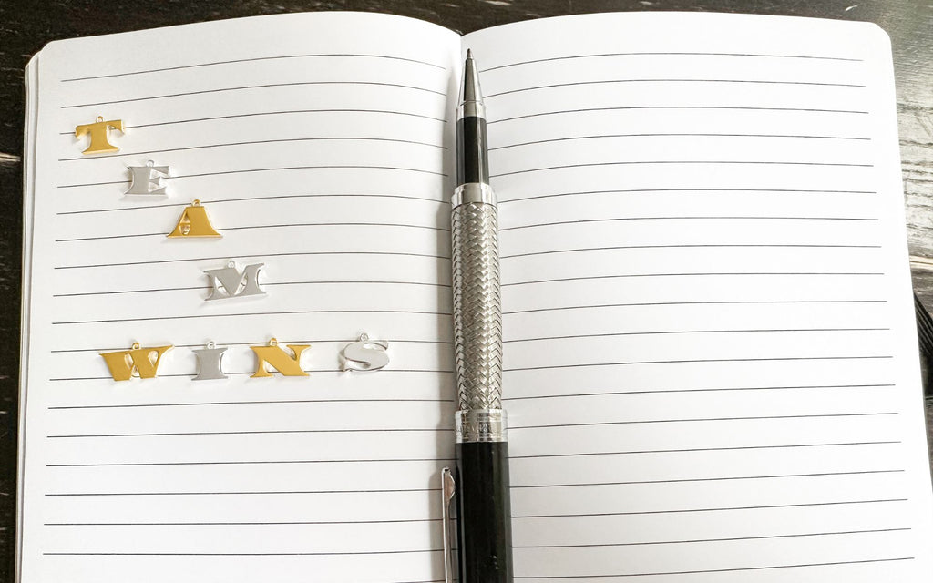 A Notebook with a pen in the middle. In silver and gold letter necklaces, Team Wins is spelt out.