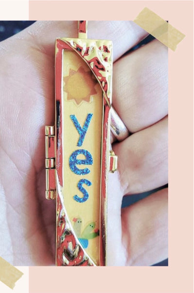 Shown Here: Julie's hand holding a locket with a yellow fortune, featuring a sun and the word "Yes" in blue.