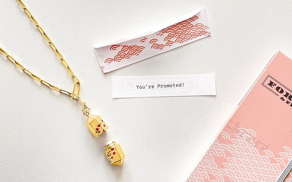 A gold Polygon Lariat Locket twisted opens and sits next to a fortune that reads "You're promoted!" Above the fortune is a fortune envelope and below it is a Fortune & Frame necklace box.