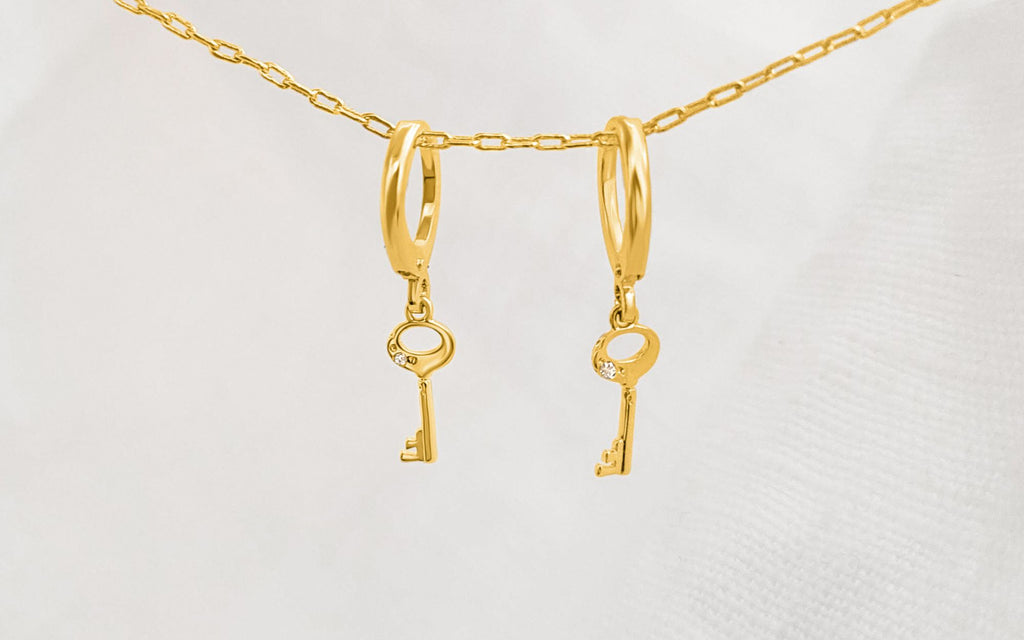 Mini gold key earrings hanging on a gold chain in front of a white cloth background.