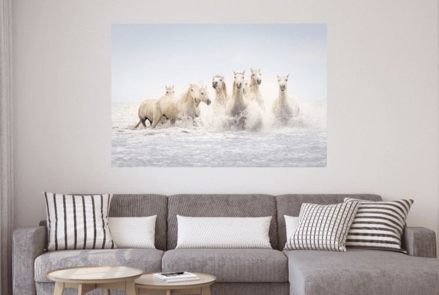 Seascape canvas wall art of white horses running in shallow ocean water on a wall above a gray couch.