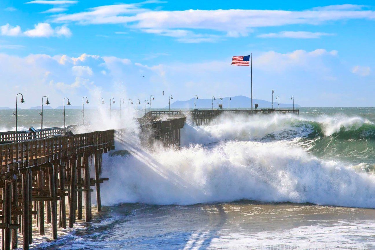 Seascape art that looks like a painting of high tide crashing into Ventura Pier in wintertime. American flag in background.