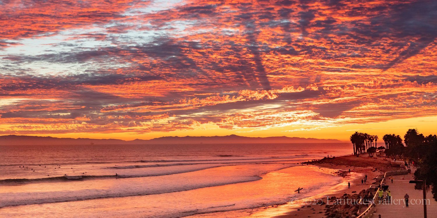 Ocean photography of surfers on the ocean beneath a bright orange sky reflected in the water.