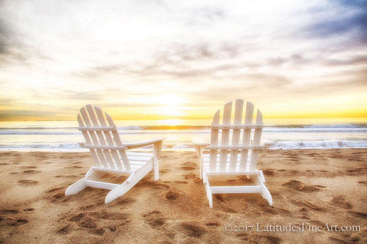 Two empty, white Adirondack chairs on a beach facing the ocean at sunset on Anacapa Island.