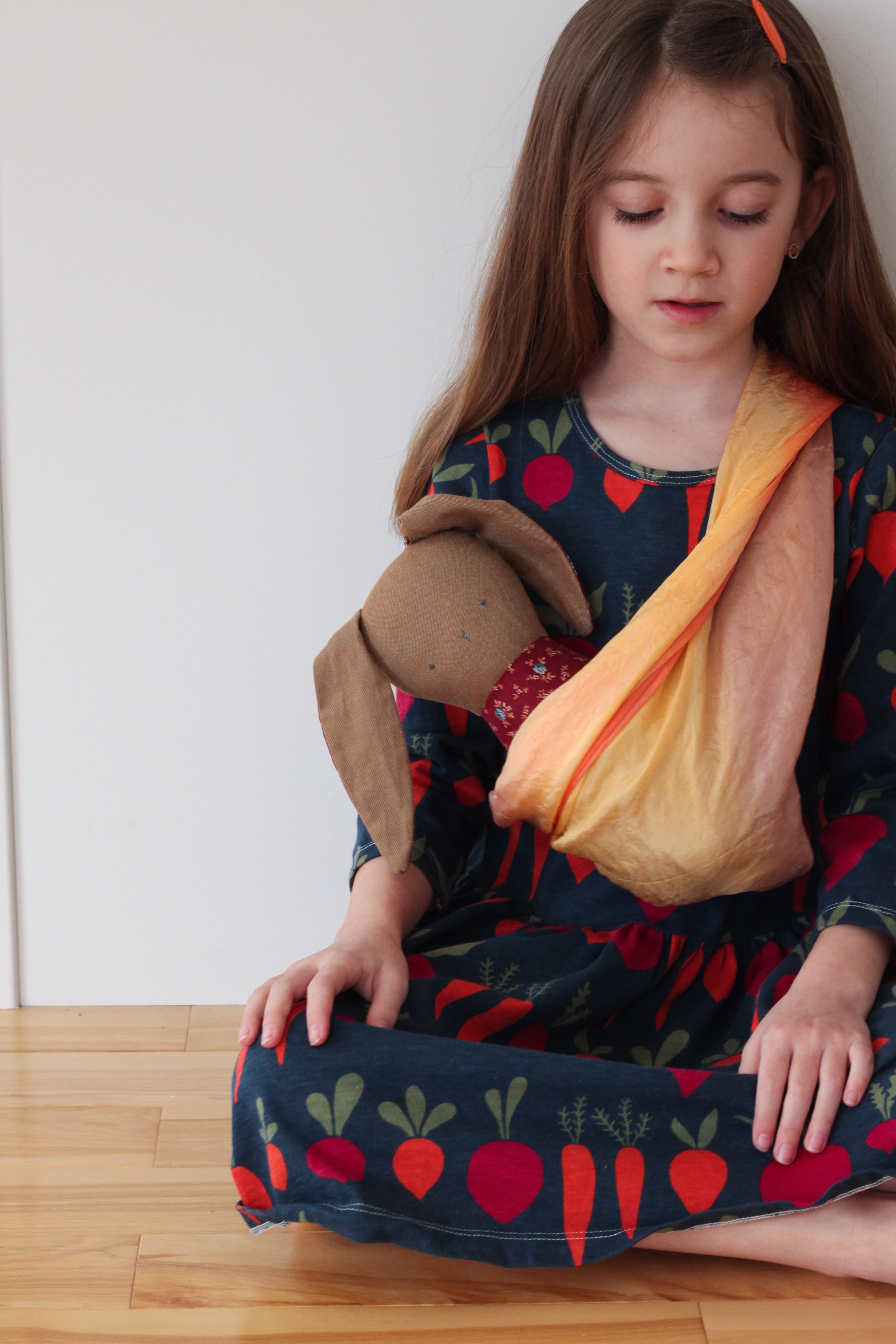 Sue's daughter is wearing a navy blue dress with orange carrots and red beets pattern. She is wearing an orange-coloured silk as a baby sling with a brown heirloom bunny doll tucked inside.