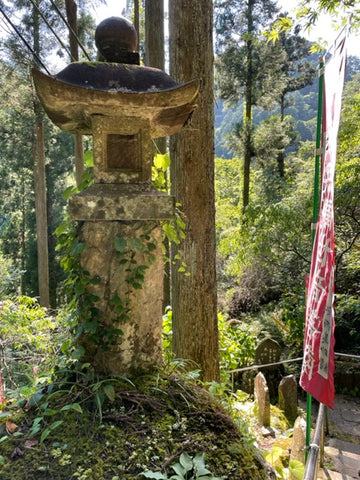 A weathered stone lantern stands amidst a lush forest setting, partially covered in moss and creeping ivy. It's situated on a mossy ground that hints at a damp and shaded environment. To the right, there is a tall, vertical red and white banner with Japanese characters, indicating a place of cultural or religious significance. The background is filled with tall, green trees, suggesting this is a serene and secluded spot, possibly within a shrine or natural park.
