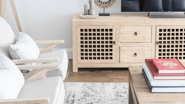 A light and airy living room featuring a natural elm wood media console with lattice-front doors and drawers. The scene includes a cosy armchair with light-coloured cushions, a rustic wooden coffee table topped with books, and a soft, patterned rug that adds texture to the space.