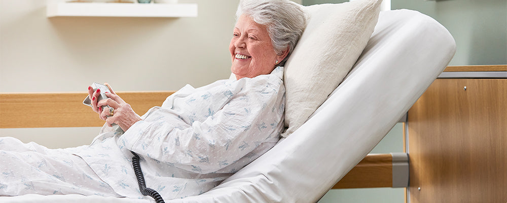 Elderly woman sat upright in a profiling care bed similing and holding a wired hand control