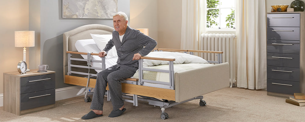 Elderly man sat on the edge of a care bed holding the rails either side of the bed