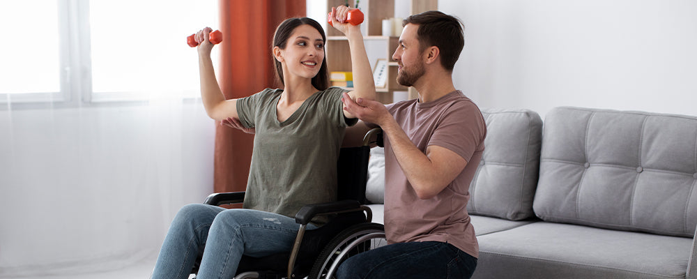 young woman with brown hair in a wheelchair holding two red hand weights above her head. Gentleman in a purple t-shirt knelt next to her support her arms 