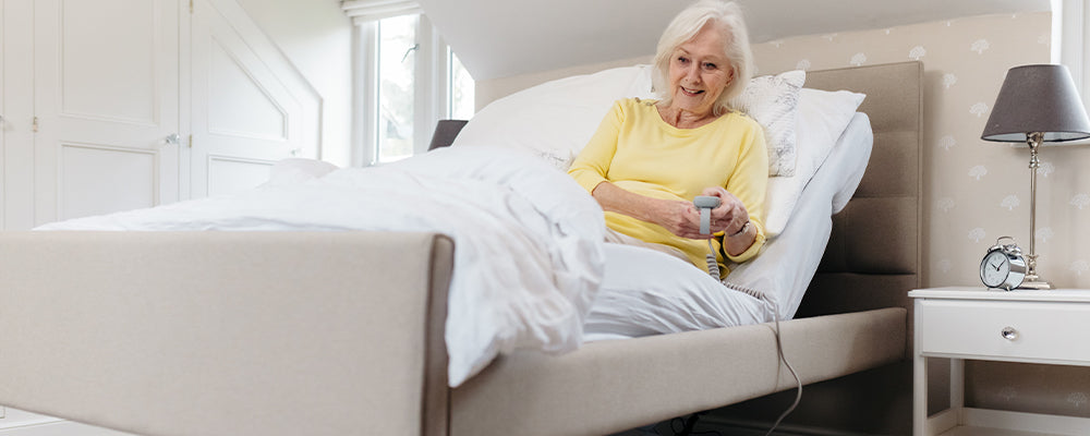 Older woman sat upright in a upholstered profiling care bed in a beige and homely bedroom