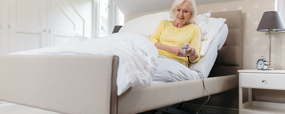 Older woman wearing a yellow top sat upright in a profiling bed and holding a remote control