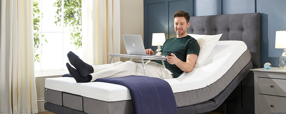A man sat upright in an adjustable bed holding a remote control with the other hand on the keyboard of a laptop