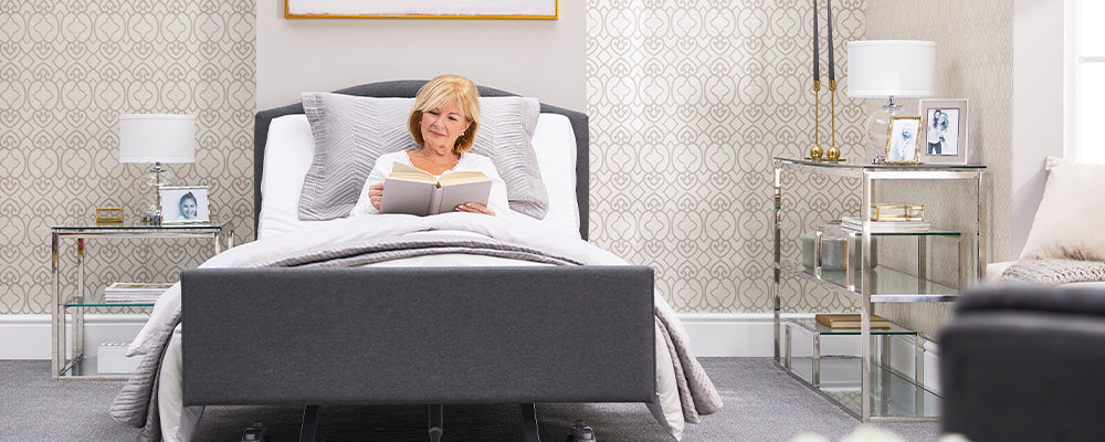 A woman with blonde hair sat upright in a profiling bed with the back rest raised up and she is reading a book