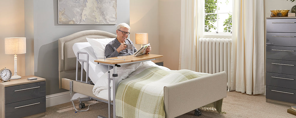 A older gentleman sat upright in a profiling bed reading a newspaper with a cup of tea