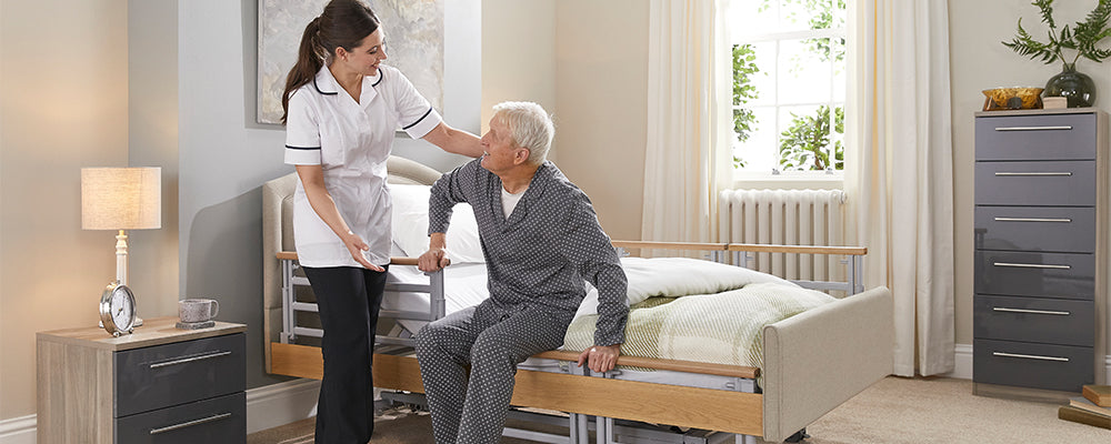 A woman with brown hair in a white tunic assisting an elderly gentleman out of bed