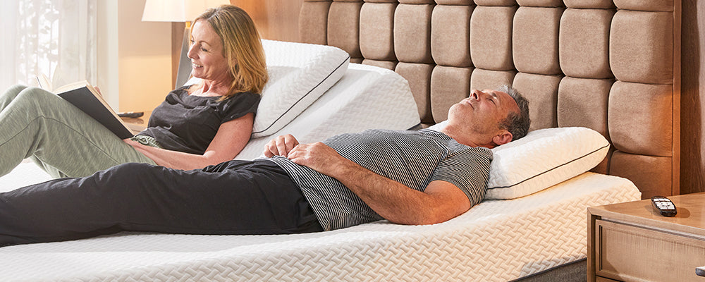 A couple in a dual adjustable bed, the woman is reading a book and the male is alseep