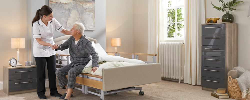 An elderly gentleman sat at the end of a bed holding his carers hand smiling