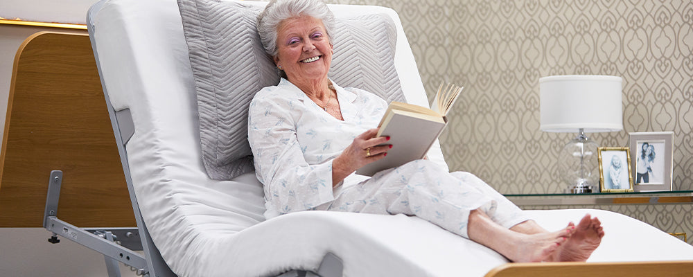 Woman sat upright in the forward tilt position of a profiling bed sat smiling holding a book
