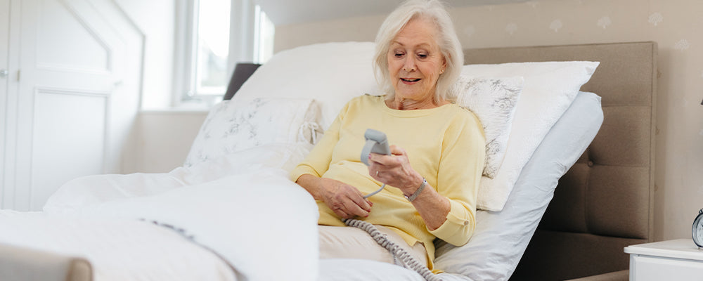 Elderly woman wearing a yellow jumper sat upright in a profiling bed holding the wired remote control